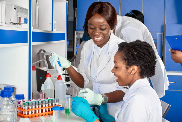 Εγκαταστάσεις δοκιμών. Νεαρές Αφρικάνες επιστήμονες ή φοιτήτριες ιατρικής, θετικές ενεργητικές χαμογελαστές γυναίκες που γελούν εργάζονται μαζί εξετάζοντας το αίμα, το νουκλεϊκό οξύ και άλλες ιατρικές εξετάσεις για μολύνσεις από ιούς. - Φωτογραφία, εικόνα