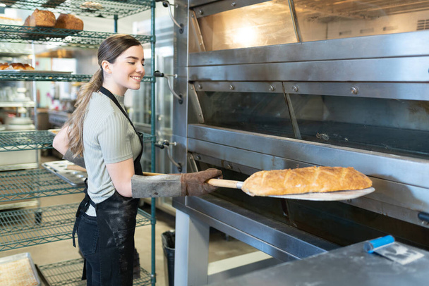 Happy female baker using a peel to take out a loaf of bread of the oven in a bakery - 写真・画像