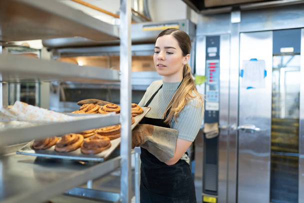 Gut aussehender junger Bäcker stellt in einer Bäckerei ein heißes Blech mit gebackenem Brot im Kühlregal auf - Foto, Bild