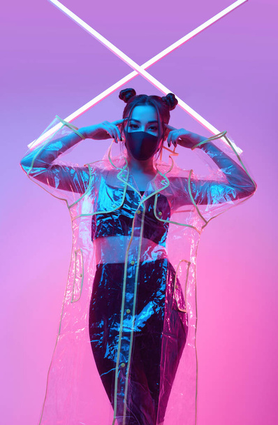 Portret van een prachtig virus masker Aziatische vrouw dragen gezichtsbescherming en regenjas rond kleurrijke heldere neon uv lichten poseren in studio - Foto, afbeelding