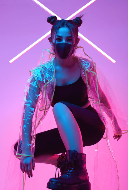 Portrait of beautiful virus mask Asian woman wearing face protection and raincoat around colourful bright neon uv lights posing in studio - 写真・画像