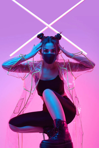 Portrait of beautiful virus mask Asian woman wearing face protection and raincoat around colourful bright neon uv lights posing in studio - Photo, image