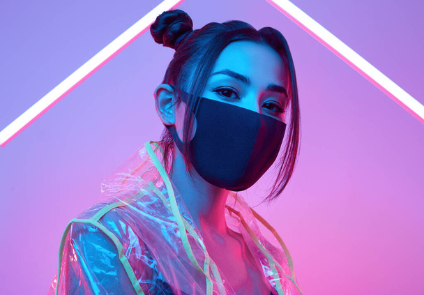 Portrait of beautiful virus mask Asian woman wearing face protection and raincoat around colourful bright neon uv lights posing in studio - Photo, Image