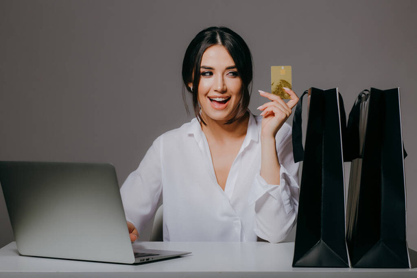 Una chica hermosa compradora satisfecha en una camisa blanca, con un ordenador portátil, y con una tarjeta de crédito en las manos, preparándose para el viernes negro aislado en gris
 - Foto, imagen