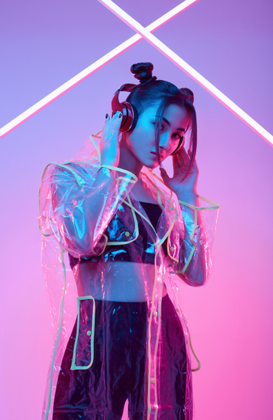 Retrato de elegante hermosa mujer asiática en un impermeable de moda escucha música en los auriculares alrededor de luces de neón brillante colorido posando en el estudio
 - Foto, imagen