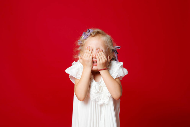 Happy young blonde curly model girl hiding her face with hands in white dress isolated on red. Little girl playing hide and seek - Photo, Image