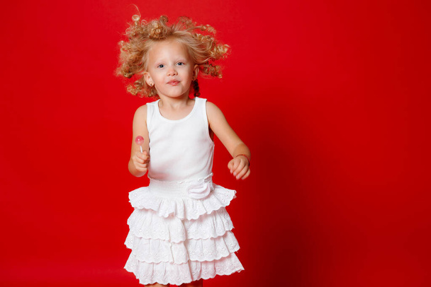 Hermosa niña rubia rizada en un vestido blanco comiendo piruleta saltando aislado en rojo
 - Foto, Imagen