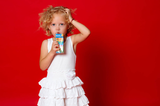 Menina encaracolado bonito no vestido branco beber suco isolado no fundo vermelho
 - Foto, Imagem