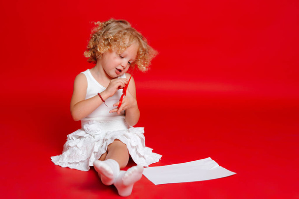Cute blonde curly girl cutting paper with red scissors isolated on red - Valokuva, kuva