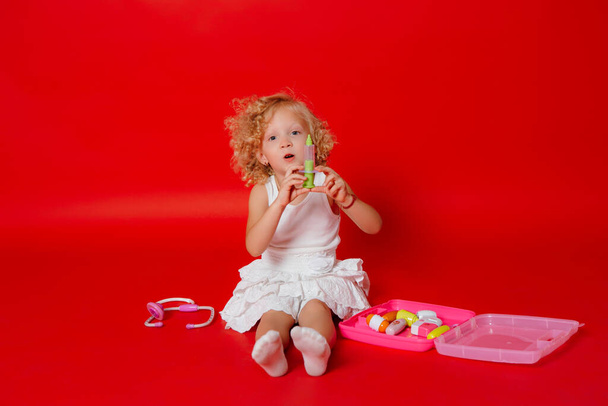 Entzückend lächelndes kleines Mädchen, das mit einer roten Spielzeugspritze spielt. Kindheitstraum Arzt zu werden - Foto, Bild