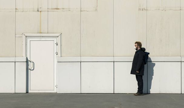 man in mask lonely on street profile portrait, isolation conditions depressive minimalism style urban photography on white exterior wall and door background with empty copy space for your text here - Foto, immagini