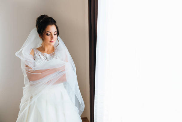 Gyönyörű menyasszony menyasszonyi ruhában és menyasszonyi fátyol öleli magát. menyasszony áll az ablaknál - Fotó, kép