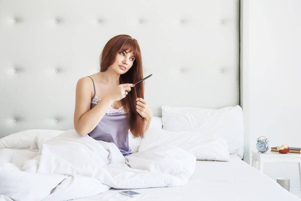 Mulher bonita penteando seu longo cabelo castanho em um quarto branco sentado na cama. Bom dia. Cuidados com o cabelo
 - Foto, Imagem