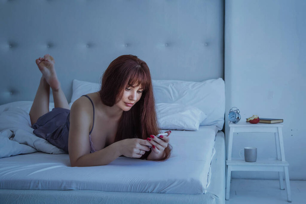 Концепція спілкування та способу життя - щаслива молода жінка лежить у ліжку зі смартфоном у домашній спальні. Сіра шовкова піжама. Ніч, сутінки
. - Фото, зображення