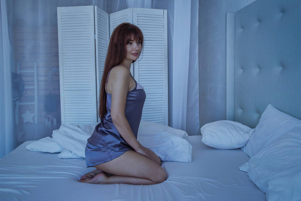 Beautiful woman sitting on a bed in silk gray pajamas. White interior. Goodnight. - Фото, зображення