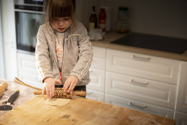Child girl, 3 years old, in white kitchen, flattening pizza dough with a small rolling pin on a wooden board.  Lockdown activity idea for preschoolers. - Photo, Image