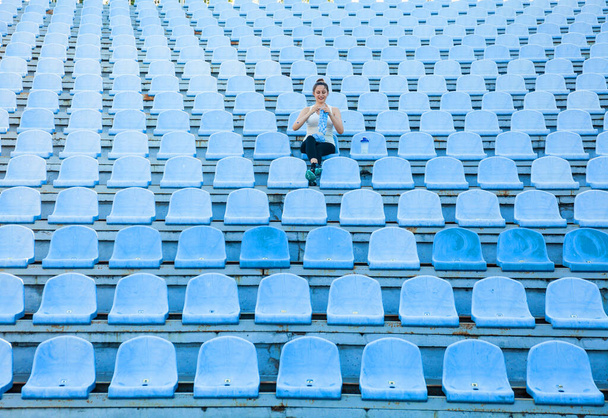 Eine junge dunkelhaarige Frau im Sportanzug im leeren Stadion strickt einen blauen Pullover. Überall kreativ werden - Foto, Bild