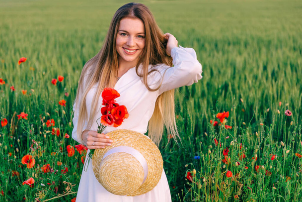 young woman with long hair looks into the camera on the background of the field. girl holding boater and bouquet of poppies.  - Photo, Image