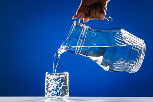 Hand pouring water from glass jug to glass isolated on a blue background - Foto, afbeelding