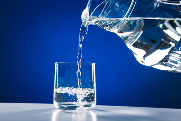 Verter agua de la jarra de vidrio al vidrio aislado sobre un fondo azul
 - Foto, imagen