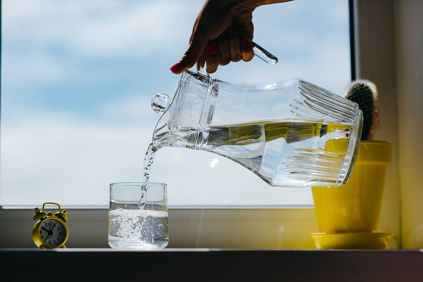 Las manos de la mujer vertiendo agua de una jarra a un vaso en un alféizar de la ventana
 - Foto, imagen