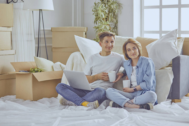 Portrét šťastného páru při pohledu na notebook spolu sedí v novém domě, obklopen krabicemi - Fotografie, Obrázek