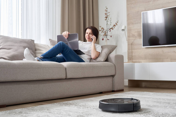 Роботизований пилосос прибирає кімнату, поки жінка відпочиває на дивані
 - Фото, зображення