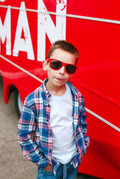 Portrait little boy wearing a checkered shirt and sunglasses in city over red background - Foto, Bild
