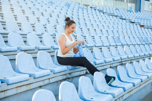 Młoda ciemnowłosa kobieta w sportowym garniturze na pustym stadionie robiąca na drutach niebieski sweter. Zdobądź kreatywność wszędzie - Zdjęcie, obraz