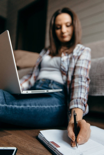 Kumral kadın dizüstü bilgisayarla yerde oturuyor ve sol elini not alıyor. İç kısmı gri. Ev ödevi serbest çalışan kadın.. - Fotoğraf, Görsel