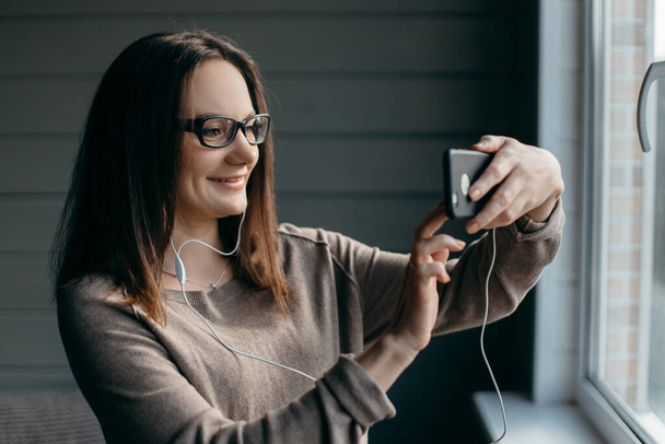 Szczęśliwa brunetka w okularach robi facetime wideo rozmowy ze smartfonem w domu, przy użyciu zoom meeting aplikacja online, odległość społeczna, praca z domu, praca zdalnie koncepcja. kwarantanna. - Zdjęcie, obraz