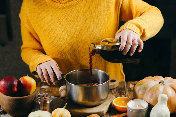 A young woman in a warm, knitted, yelow pullover is pouring wine from glass bottle to a pan to make hot mulled wine with spices and citrus fruits. A cozy, winter evening. Winter drinks. - Фото, изображение