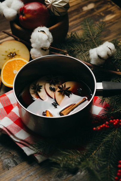 Mulled wine hot drink with citrus, apple and spices in aluminum pot on checked tablecloth. Winter holiday mood with fur tree branches, red berries and cotton. - Photo, image