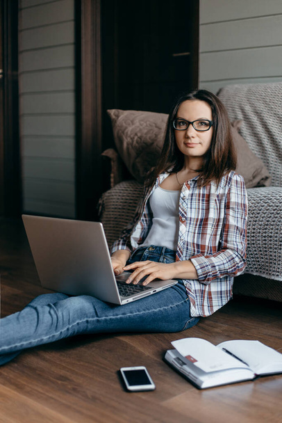Young  brunette woman in glasses sitting  on the floor, working with laptop at home. Look at camera. Homework freelancer woman - Working home concept  - Photo, Image