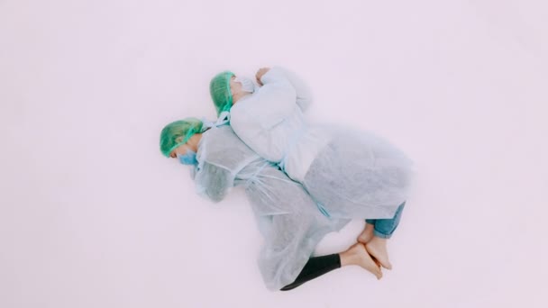 Két nő védőruhában és arcmaszkban. Fiatal lányok fekszenek a fehér padlón védőruhában, sapkában és maszkban. Az influenzavírus világjárvány tragédiája. - Felvétel, videó