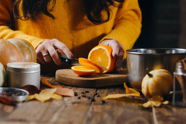 Close-up image of manicured woman's hands cutting oranges on wooden board for hot wine. Pumpkins, leaves, candles on the table. Autumn mod. - Foto, Imagen