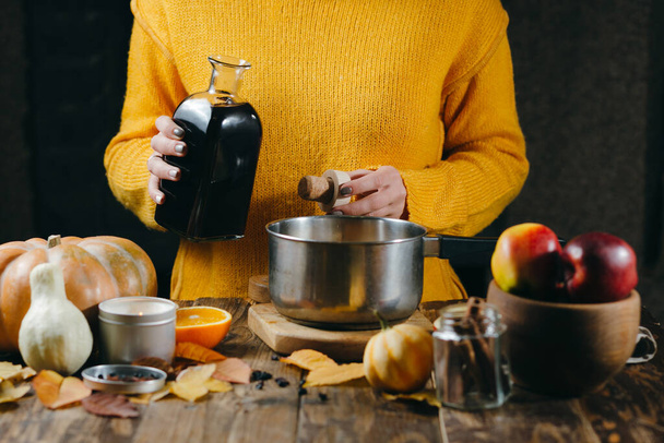 A young woman in a warm, knitted, yelow pullover is pouring wine from glass bottle to a pan to make hot mulled wine with spices and citrus fruits. A cozy, winter evening. Winter drinks. - Valokuva, kuva