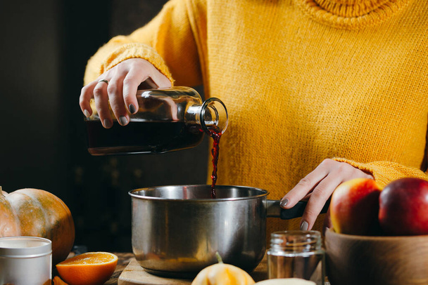 Een jonge vrouw in een warme gebreide trui giet wijn uit een glazen fles in een pan om warme glühwein te maken met specerijen en citrusvruchten. Een gezellige, winteravond. Winterdrankjes. - Foto, afbeelding