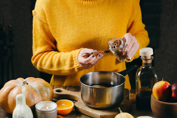Подготовка глинтвейна Руки женщины в желтом свитере добавляют палочки корицы к горячему глинтвейну
. - Фото, изображение
