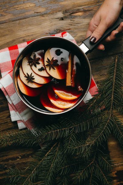 Woman's hand holding a pot with hot mulled wine with citrus, apple and spices in aluminum pot on checked tablecloth. Winter holiday mood with fur tree branches. - Foto, immagini