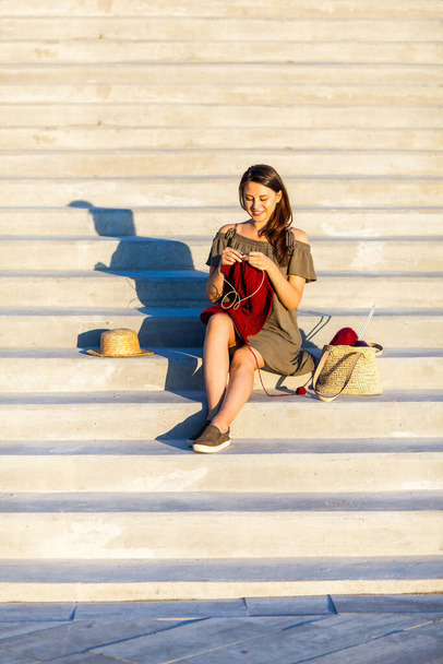 A young woman sitting In a deserted place cityscape, on the steps and knitting a sweater with knitting needles on a summer day. Get Creative everywhere during quarantine - Photo, image