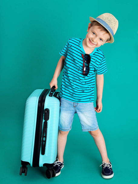 The boy in the hat picks up a heavy blue suitcase and travels on a colored background - Фото, изображение