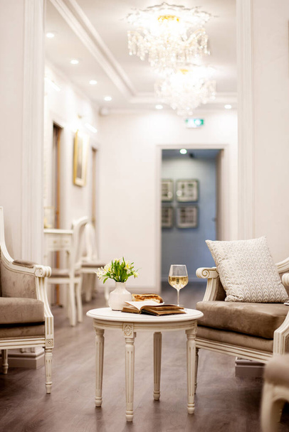 Een vaas met mooie witte bloemen, een glas wijn en knapperig gebak staan op tafel in een lichte en gezellige kamer. Schieten in het interieur - Foto, afbeelding