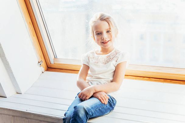Restez à la maison Restez en sécurité. Petite fille douce et mignonne souriante en jeans et T-shirt blanc assis sur le rebord de la fenêtre dans le salon lumineux à la maison à l'intérieur. Enfance écoliers jeunesse relax concept - Photo, image
