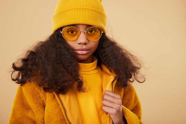 Stylish African American preteen girl with curly hair wearing yellow fur jacket and hat with glasses looking at camera against beige background - Foto, imagen