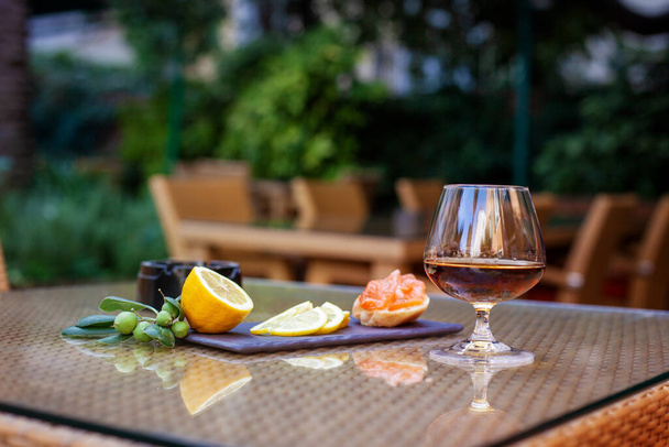A glass of cognac, slices of lemon, a sandwich and a sprig of olive lie on the table on the restaurant's terrace. Rest, relaxation - Foto, imagen