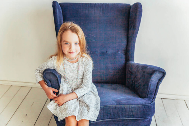 Stay Home Stay Safe. Sweet little girl in white dress sitting on modern cozy blue chair relaxing in white bright living room at home indoors. Childhood schoolchildren youth relax concept - Fotografie, Obrázek