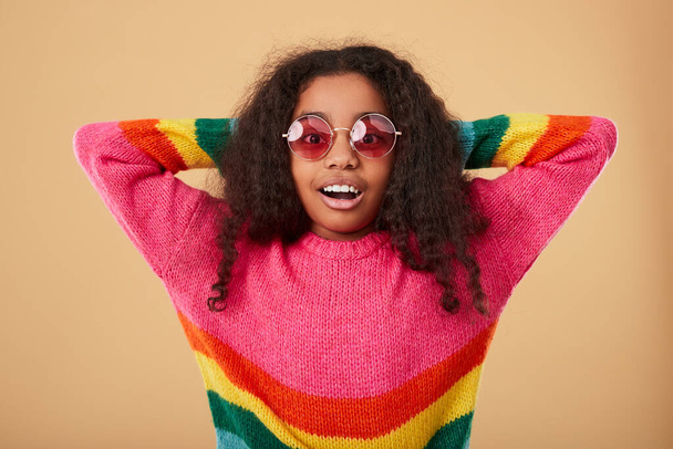 Happy amazed African American girl in trendy sunglasses and colorful striped sweater looking at camera against beige background - Foto, immagini