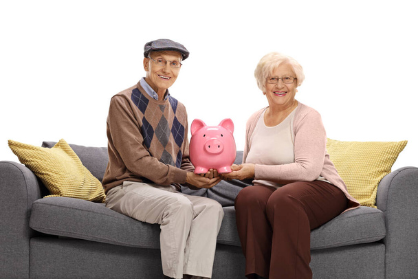 Elderly man and woman sitting on a sofa and holding a piggy bank isolated on white background - Фото, изображение