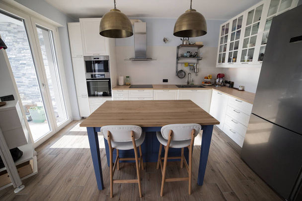 Weiße, helle Küche mit blauer Insel und doppelten Messing-Pendelleuchten. Zentrale Sicht. - Foto, Bild